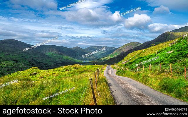 Long country road leading to majestic Black Valley with lake and mountain range covered in sunlight and shadows from clouds, County Kerry, Ireland