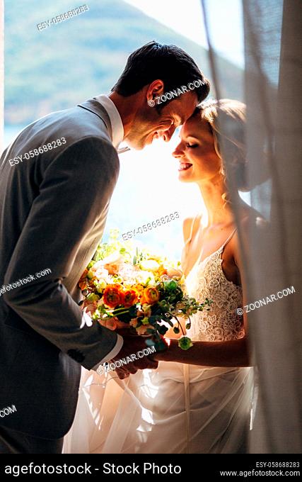 The bride and groom are sitting at the window hugging each other, smiling and holding each other's hands. In their hands a bridal bouquet