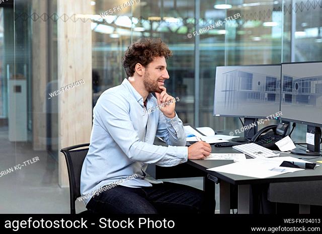 Thoughtful male entrepreneur writing while sitting at computer desk in factory