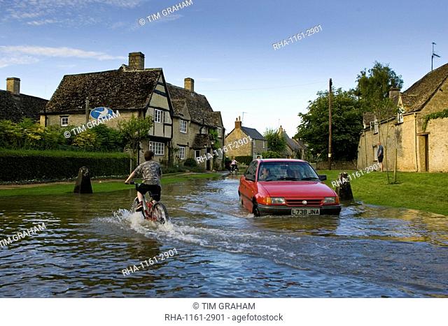 Car and boy on a bike try to move through flood water in Minster Lovell, Oxfordshire, England, United Kingdom