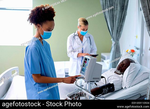Mixed race female doctor wearing face mask using blood pressure monitor in patient room