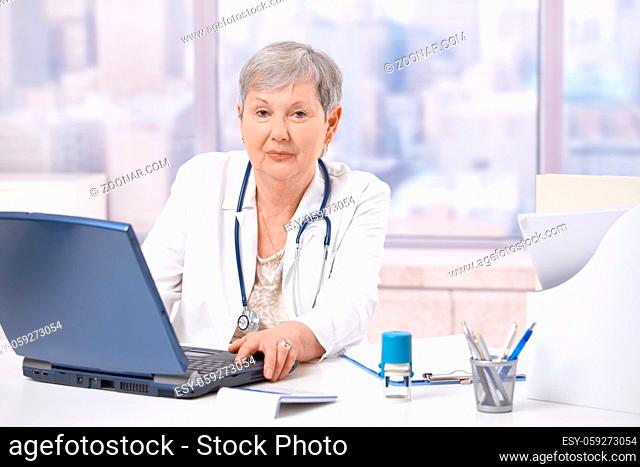Senior female doctor, working at desk, using laptop computer. Looking at camera