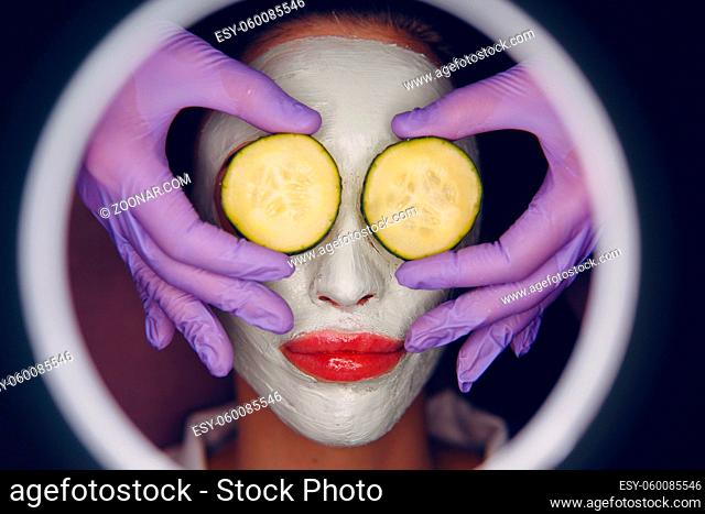 Young beautiful woman receiving facial clay mask with cucumber on her eyes in spa beauty salon with magnifying glass