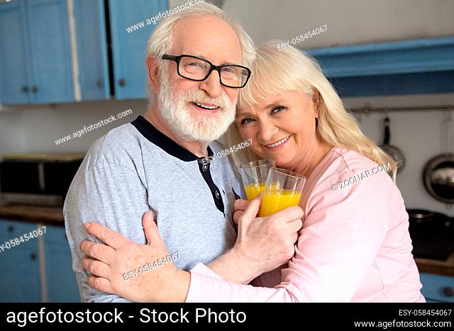 Portrait of two sweet elderly people smiling in kitchen. Senior couple hugging and holding glasses with orange juice at home