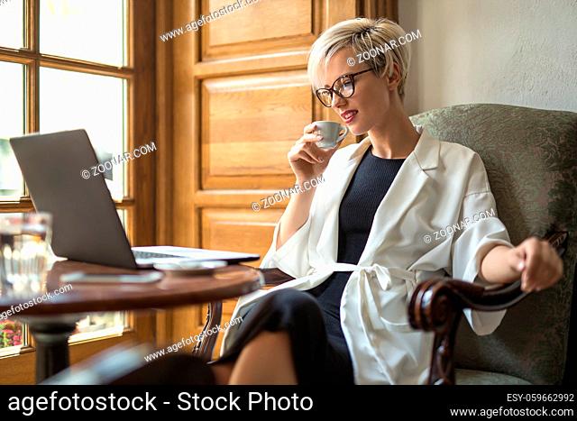 Charming blonde girl with a smile in glasses sits on the armchair at the table in the restaurant. She holds a cup in right hand and looks at the laptop