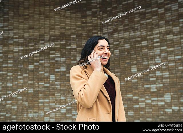 Happy young woman talking on smart phone in front of wall