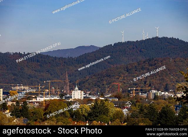 27 October 2021, Baden-Wuerttemberg, Freiburg: Wind turbines stand on a foothill of the Black Forest while the city of Freiburg with the tower of the Freiburg...