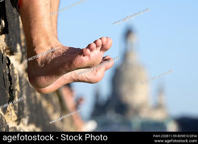 11 August 2023, Saxony, Dresden: Passers-by sit on a wall on the banks of the Elbe in front of the backdrop of the Frauenkirche and let their feet dangle