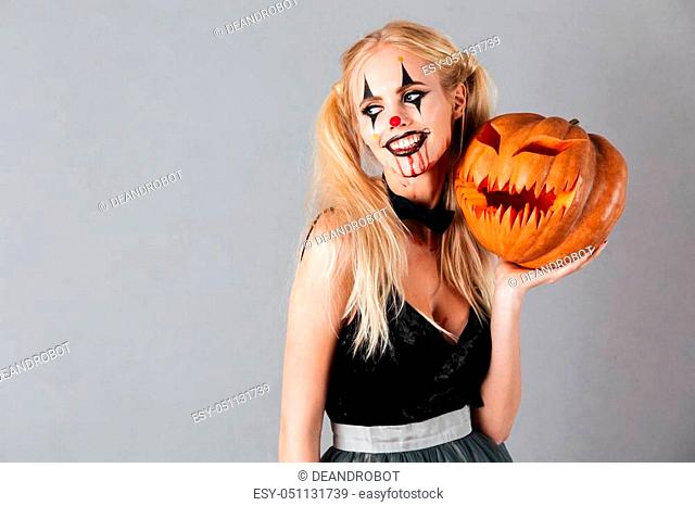 Smiling blonde woman in halloween make up posing with carved pumpkin and looking away over gray background