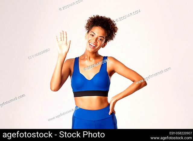 Concept of sport and workout. Attractive, healthy african-american female athlete, waving hand friendly to say hello, greeting someone, white background