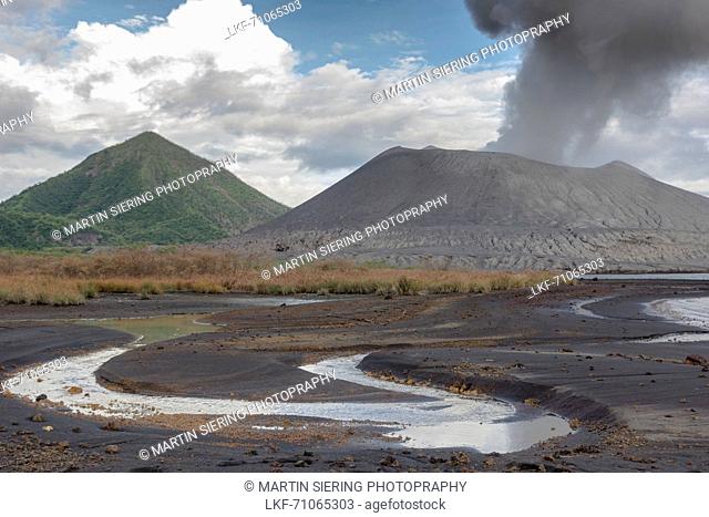 Eruption of the active volcano Tavurvur with ash cloud. In the foreground rainwater in the ash sand, in the background the green cone of the „Vulcan' volcano