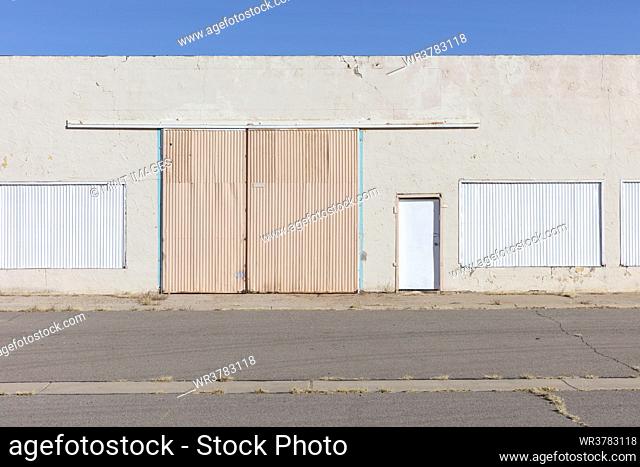 Closed warehouse building with metalwork shutters on doors and windows, and weeds growing through tarmac