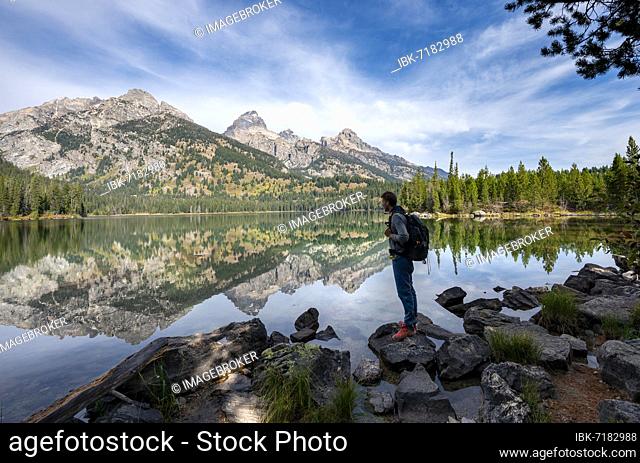 Young man standing by a lake, reflection in Taggart Lake, view of Teton Range mountain, peaks Grand Teton and Teewinot Mountain, Grand Teton National Park
