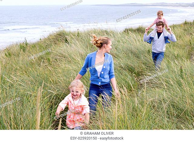 A young family of four running through long grass next to the beach. They are wearing casual clothing and are smiling