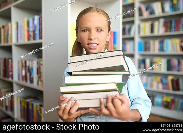 Waist-up portrait of a sad teenage girl with a pile of books standing in the school library