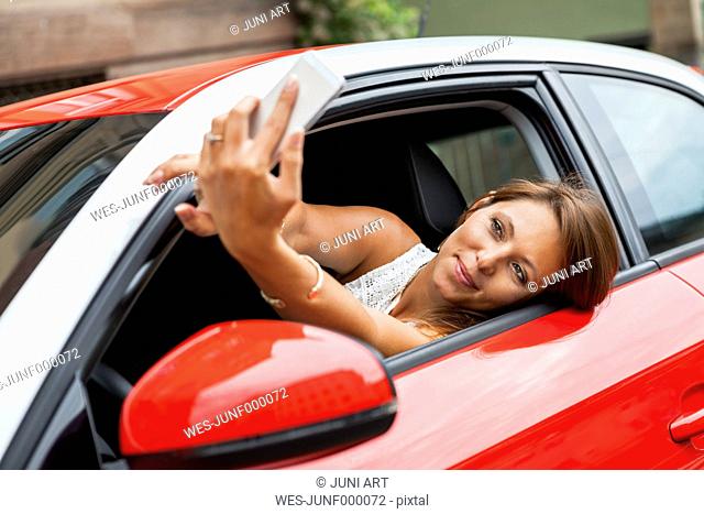 Portrait of smiling young woman sitting in her car taking a selfie with her smartphone