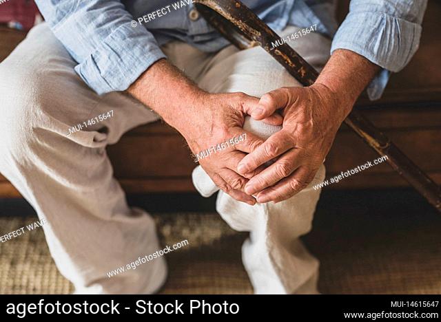 Close up of senior old man facing knee problem, sitting on sofa holding knee at home. Elderly man suffering from severe knee ache sitting with walking stick in...