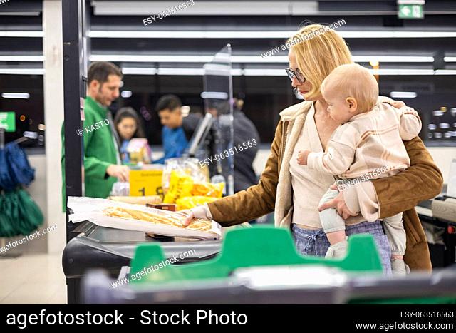 Mother shopping with her infant baby boy, holding the child while stacking products at the cash register in supermarket grocery store