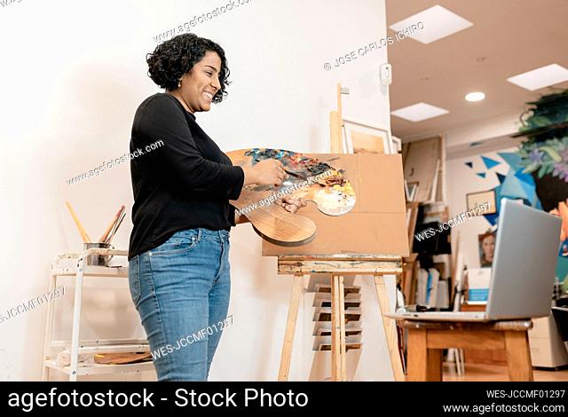 Smiling female artist on video call through laptop holding palette at studio