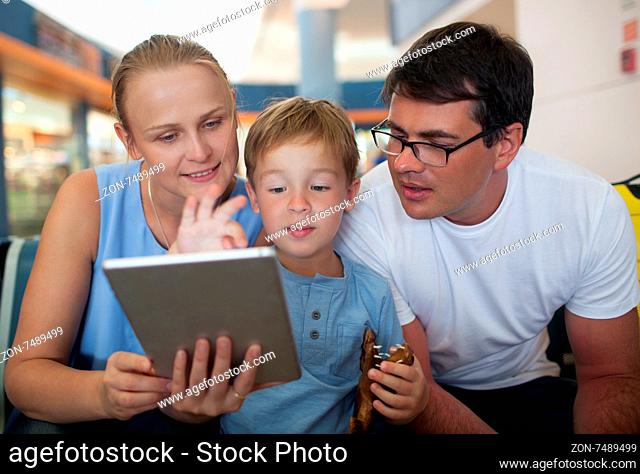 Young parents and little son at the airport. Mother holding pad and boy playing game, both parents looking at screen. Entertaining during flight waiting