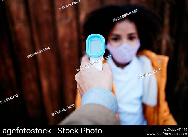 Woman using infrared thermometer while checking girl temperature against wooden wall
