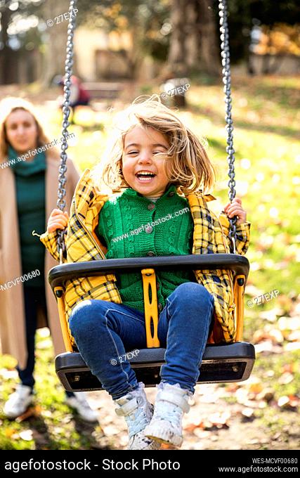 Smiling girl playing on swing with woman standing in background at park