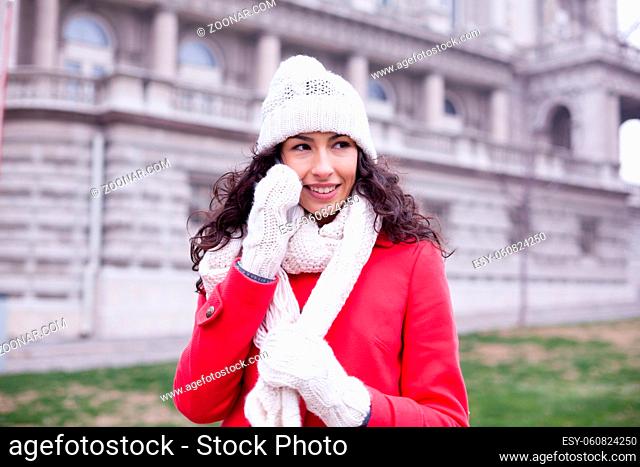 Beautiful Woman in Red Coat and and wool cap and gloves read messages on smartphone and smiling. Urban Space. Cold Weather