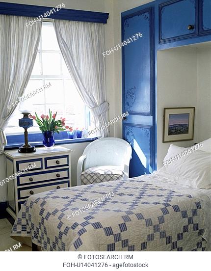 Blue+white patchwork quilt on bed in alcove with fitted blue cupboards in bedroom with white curtains