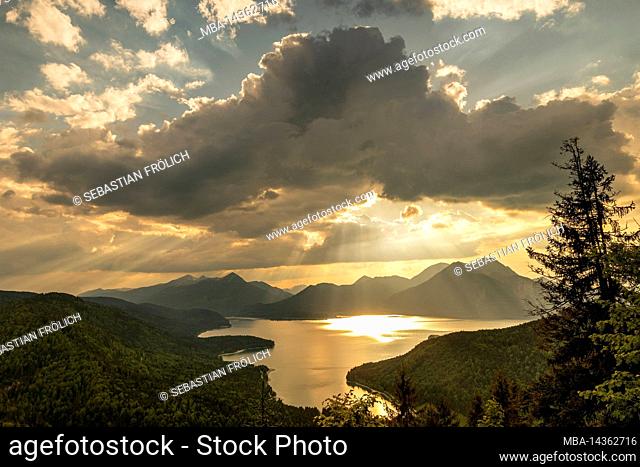 Sun rays and golden light over Walchensee, with Herzogstand, Heimgarten and Simetsberg in the background. The lake reflects the color and dense clouds in the...
