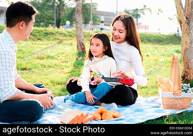 Happy Asian young family father, mother and children having fun and enjoying outdoor together sitting on the grass party with playing Ukulele during a picnic in...