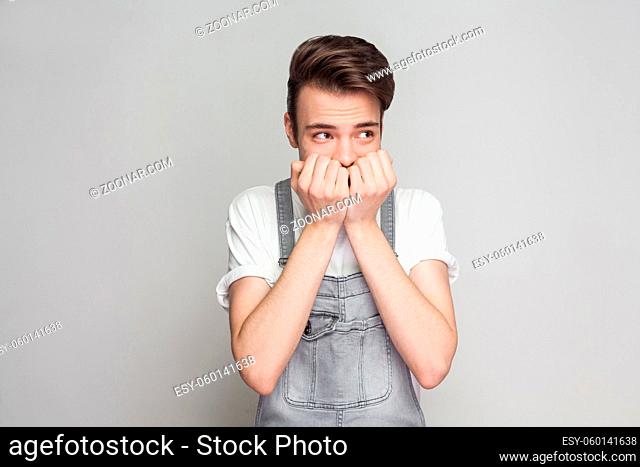 Portrait of nervous young brunette man in casual style with white t-shirt and denim overalls standing and looking away, biting nails with worry face