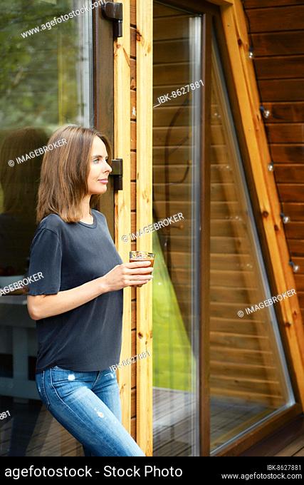 Cute woman came out of a wooden house, leaned against a glass door and drinks juice from a glass