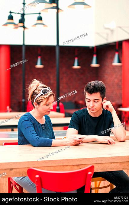 Woman and young man talking together and using mobile phones sitting by a table in cafe
