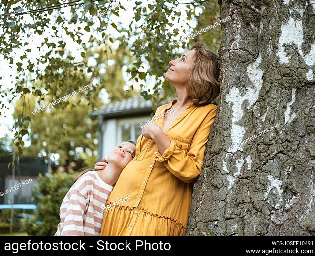 Smiling mother with daughter leaning on tree trunk