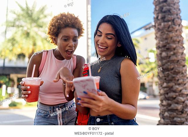 USA, Florida, Miami Beach, two happy female friends with cell phone and soft drink in the city