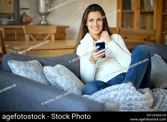 beautiful young smiling woman in white sweater texting on her phone on a gray sofa