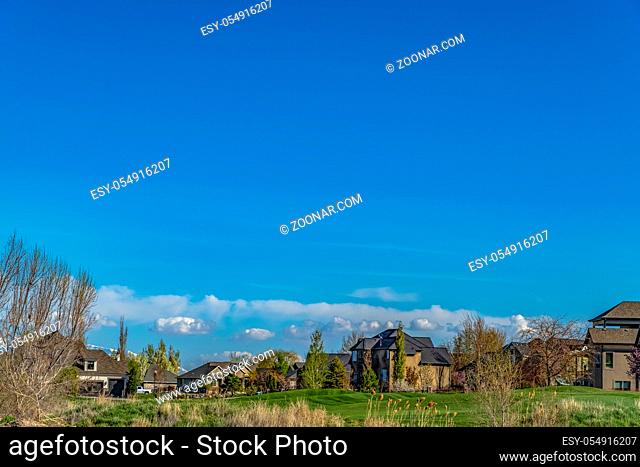 Houses on a rich green field with trees and brown grasses in the foreground. Brilliant blue sky with puffy clouds can be seen in the background on this sunny...