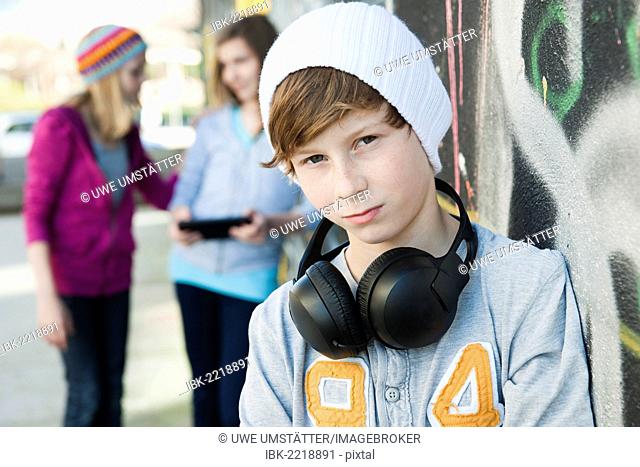Boy wearing headphones standing in front of a wall with graffiti, with two girls behind him