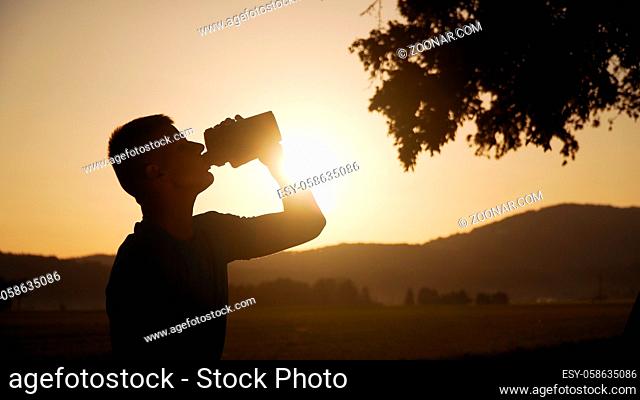 silhouette of fit young man drinking water with sun and nature behind it in sunset