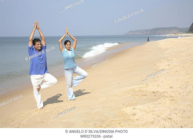 South Asian Indian young man and woman doing yoga standing on one folded legs and raising joint hand palms overhead on seashore , Shiroda , Dist Sindhudurga