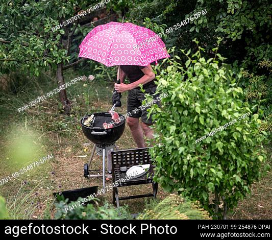 01 July 2023, Hesse, Frankfurt/Main: A man, caught in a rainstorm, stands in his backyard at a charcoal grill, turning meat on the grate