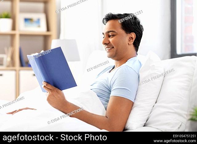 happy indian man reading book in bed at home