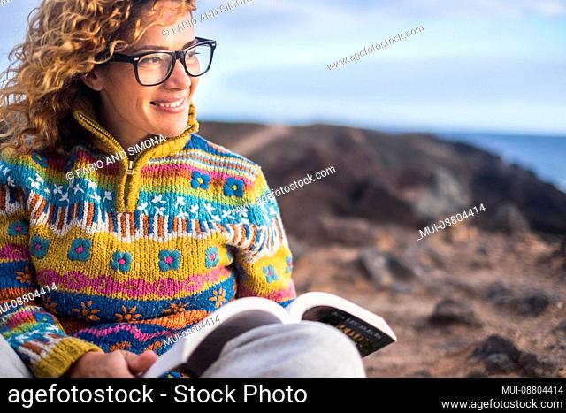 Woman sit down on the rocks in outdoor beautiful place with ocean in background reading a paper book and enjoying the freedom and the relax situation