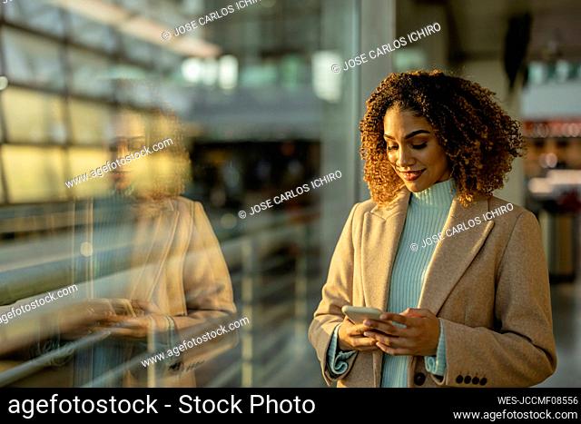 Smiling woman using smart phone by window with reflection on glass at station