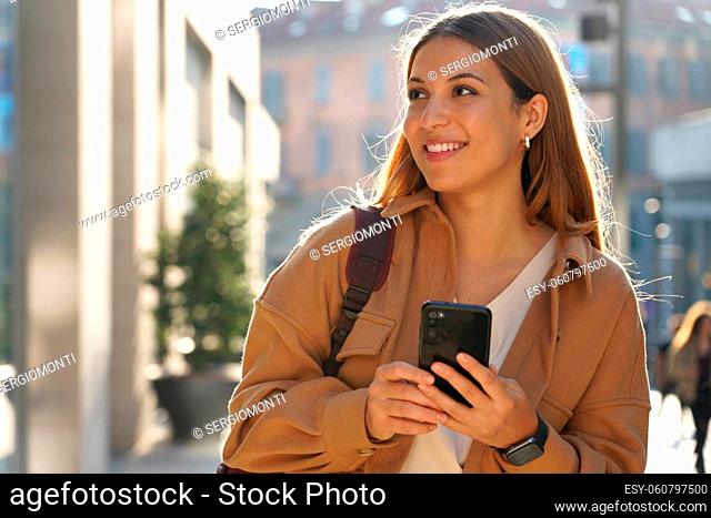 Portrait of young smiling fashion woman wearing coat holds smartphone looking through shop window on city street. Copy space
