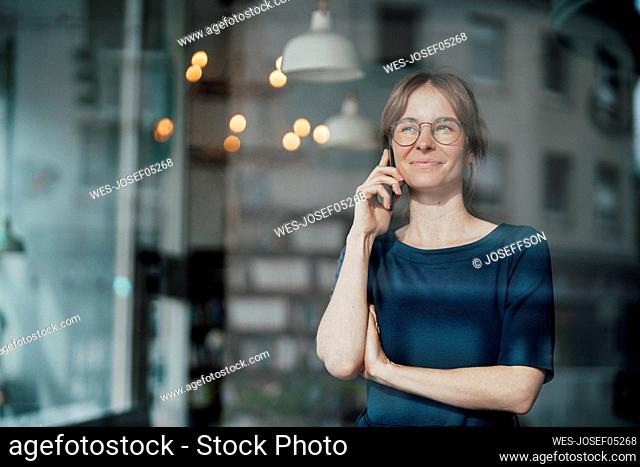 Smiling young female freelancer talking on smart phone seen through glass window