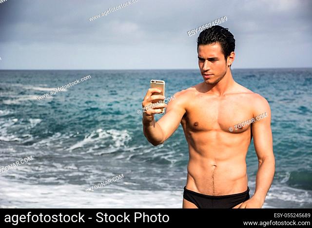 Side View of a Shirtless Young Man Taking Selfie Photos at the Beach While Standing Under the Sun