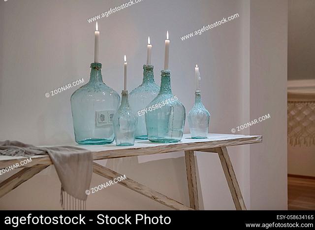 Burning candles in the stylish bottles on the wooden table on the white wall background. There is a shawl near them. Behind the bottles there are power sockets...