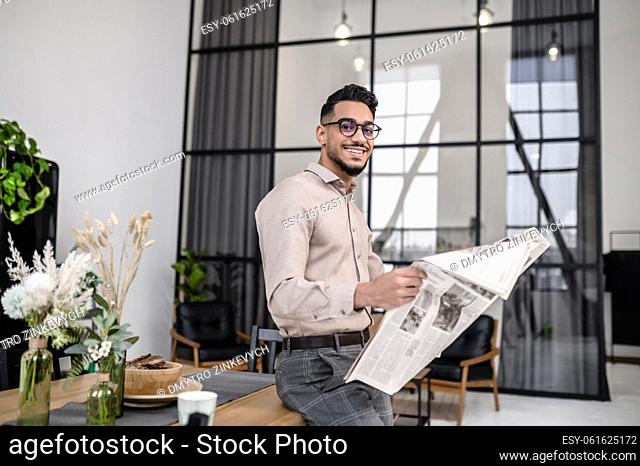 Good start. Young bearded attractive man in glasses with newspaper smiling at camera standing leaning on table in cozy bright room