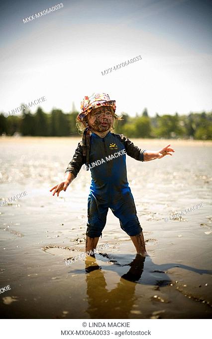 Baby boy wearing hat and blue sunsuit on standing on the beach with muddy face and arms stretched out